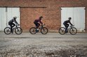 Drie fietsers rijden op een geplaveide weg. De middelste fietser draagt een bordeauxrood shirt met lange mouwen.