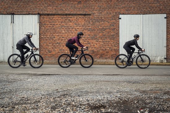 Drie fietsers rijden op een geplaveide weg. De middelste fietser draagt een bordeauxrood shirt met lange mouwen.