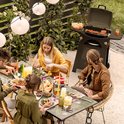 Zwarte KitchenBrothers gasbarbecue, buiten opgesteld bij een tuintafel met etende mensen. De barbecue heeft worsten en spiesjes.