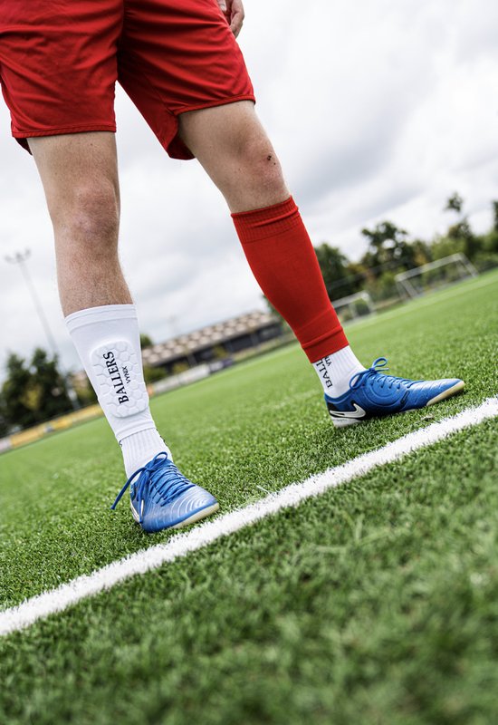 Voetballer in rood tenue met een witte Vynix Ballers scheenbeschermer aan de linkerkuit.