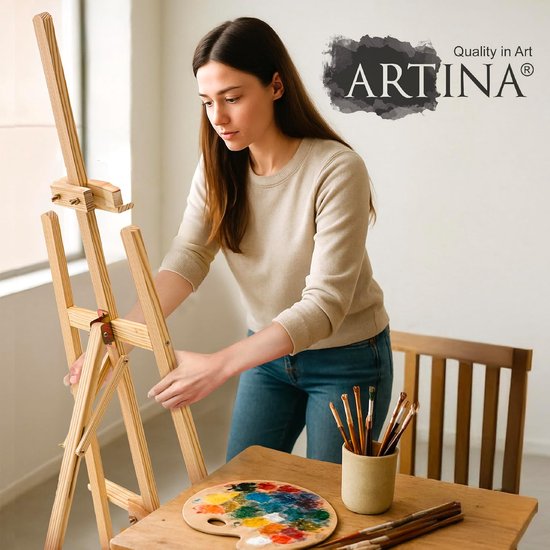 Vrouw met beige trui en spijkerbroek stelt een houten schildersezel in. Tafel met palet, verf en kwasten.