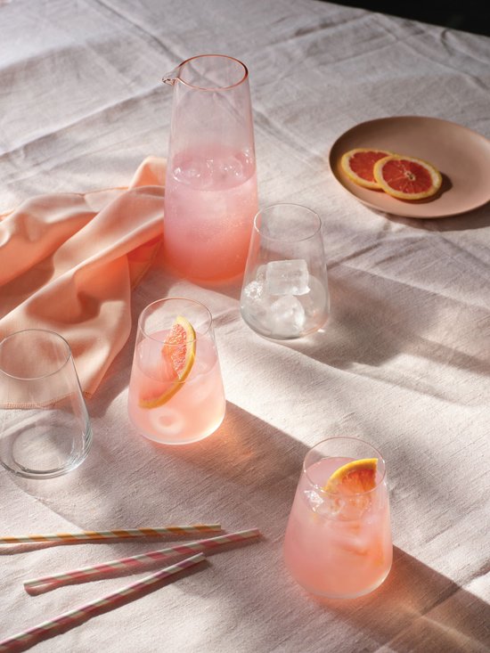 Drie glazen met een roze drankje, één glas met ijs, één leeg glas, een karaf met roze drankje, twee plakjes grapefruit en rietjes op een lichtroze doek.