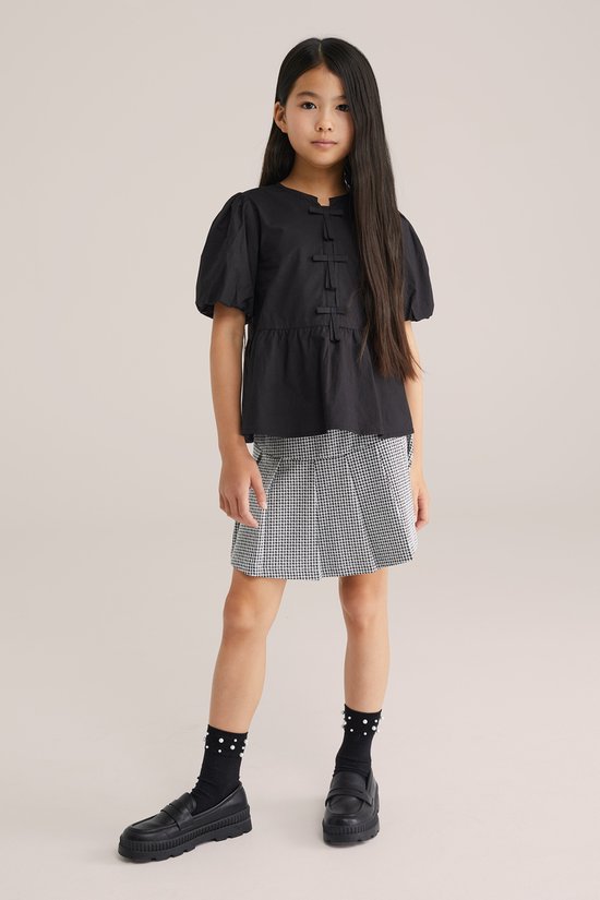 Meisje met lang haar, draagt een zwart topje en een wit-zwarte geruite rok met plooien.