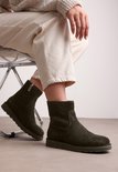 Olijfgroene suède enkellaarzen met ritssluiting, gedragen door een model zittend met beige broek.