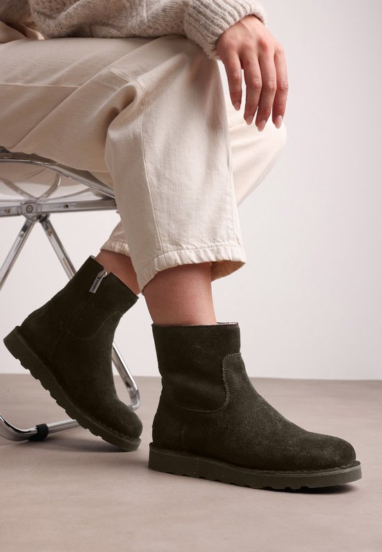 Olijfgroene suède enkellaarzen met ritssluiting, gedragen door een model zittend met beige broek.