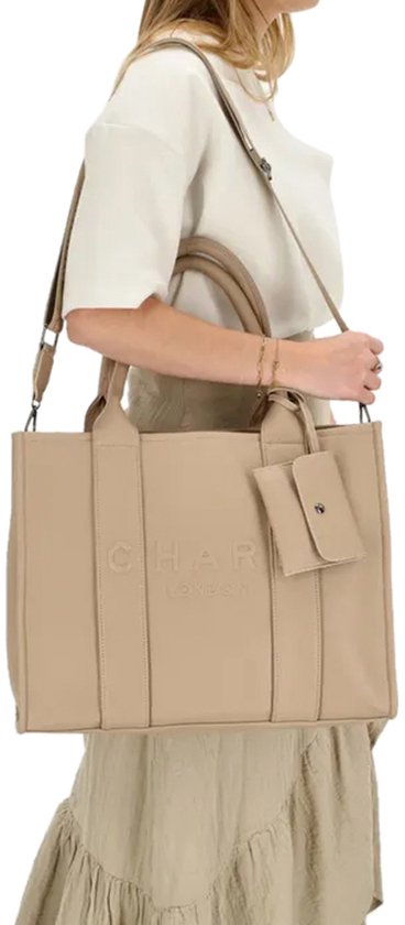 Vrouw met beige handtas met 'CHARM LONDON' logo, draagt deze crossbody met schouderband.