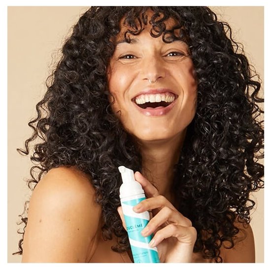 Lachende vrouw met krullend haar houdt een fles met teal- en witte spiraalvormige strepen voor droogshampoo vast.