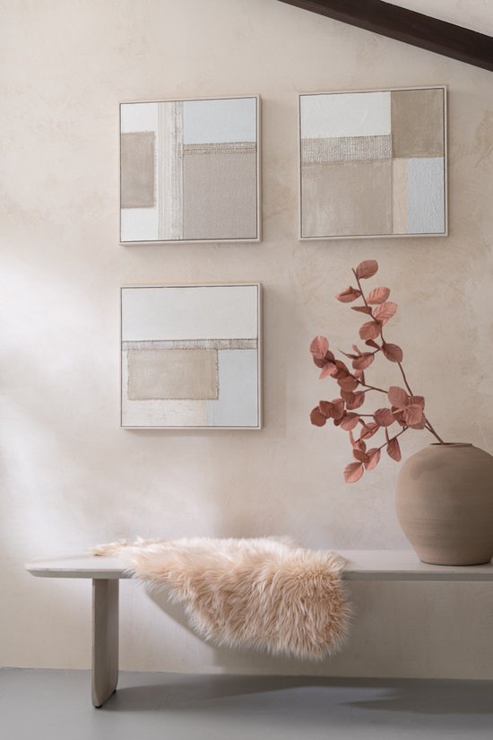 Trois peintures abstraites beiges, blanc cassé et grises, accrochées à un mur clair au-dessus d'un banc avec une fourrure.