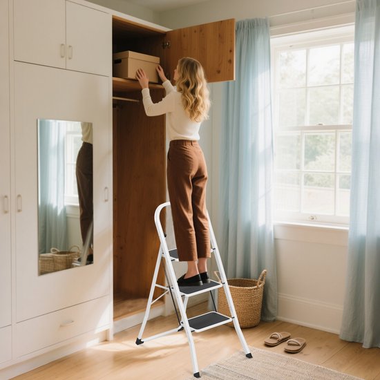 Een vrouw in een witte trui en bruine broek staat op een witte opvouwbare trap om een doos in een kledingkast te leggen.