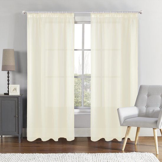 Twee crème vitragegordijnen hangend voor een raam in een woonkamer met een grijze kast en een stoel.
