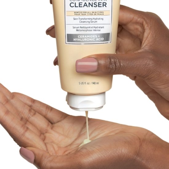 Een hand met een lichtbruine huid drukt op een tube met een beige vloeistof, die in een andere hand druppelt.