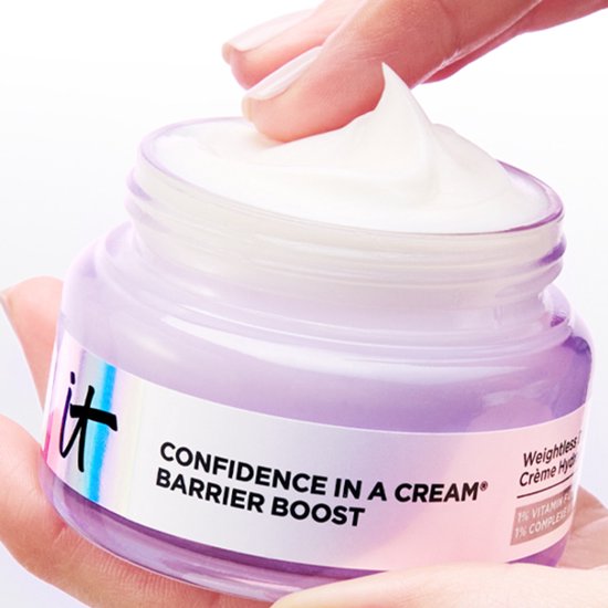 Een hand pakt een pot crème met een paars doorschijnend deksel, gevuld met witte crème.