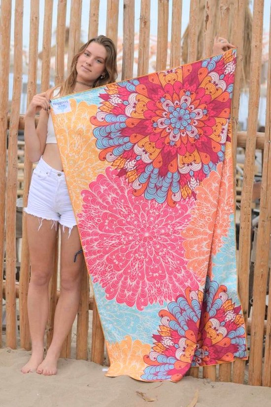 Vrouw met witte top en korte broek staat voor een houten hek en houdt een groot strandlaken met mandala print vast.