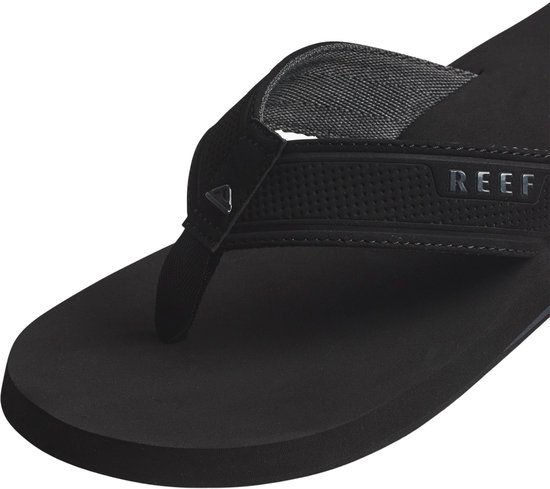 Detailopname van een zwarte Reef slipper. Focus op de band met Reef logo en textuur.