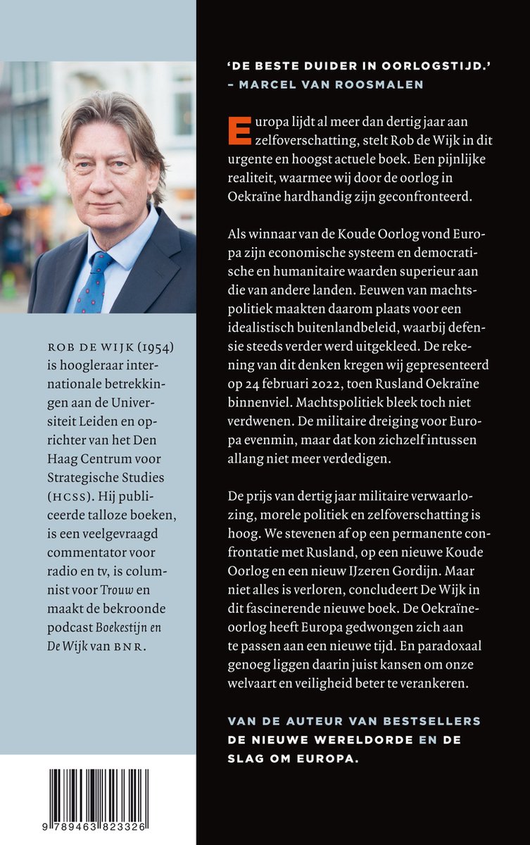 Foto van auteur Rob de Wijk, staand buiten met op de achtergrond een groen/geel gebouw. Hij draagt een blauw pak met een wit overhemd en blauwe das.