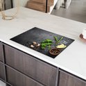 Zwarte inductie beschermer met betonprint, basilicum, rozemarijn, citroenpartjes, knoflook en peperkorrels, op een marmeren aanrecht.