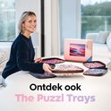 Een glimlachende vrouw zit aan tafel met puzzelbakjes en de Puzzl Trays verpakking, met de tekst 'Ontdek ook The Puzzl Trays'.