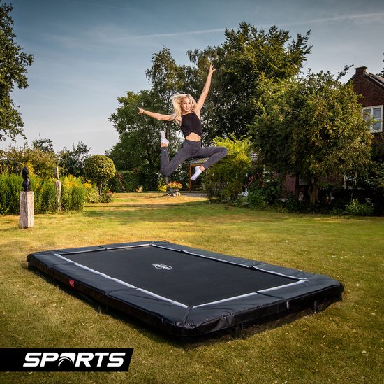 Vrouw in de lucht springend op een rechthoekige zwarte trampoline in een tuin.