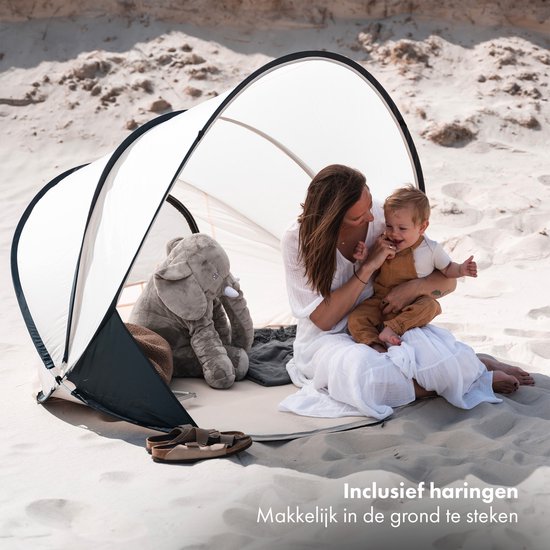 Vrouw met baby en olifantknuffel in crème strandtent met donkergrijs frame op het strand.