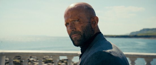 Man met geschoren hoofd, baard, en een grijs pak, kijkt naar de camera bij de zee.
