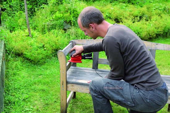 Man schuurt een houten bankje in de tuin met een rode Einhell multischuurmachine.