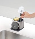 Une main place une brosse verte dans un organisateur d'évier en acier inoxydable avec éponge et savon.