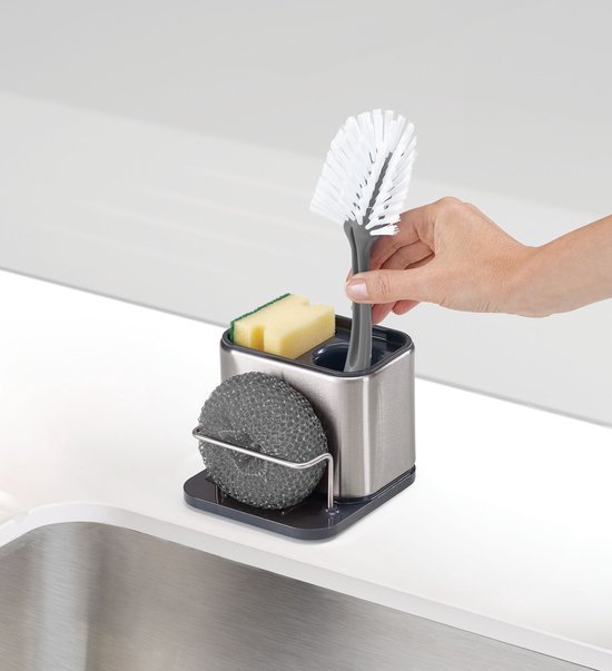 Une main place une brosse verte dans un organisateur d'évier en acier inoxydable avec éponge et savon.