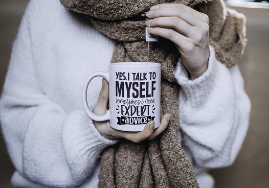 Witte mok met tekst "YES, I TALK TO MYSELF SOMETIMES I NEED EXPERT ADVICE", vastgehouden door persoon in winterkleding.