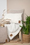 Off-white sierkussen met pomponnetjes, een gebreide plaid en decoratieve takken op een houten bankje.