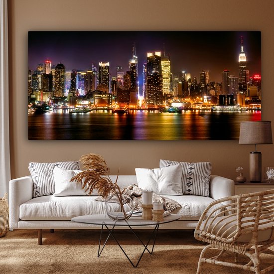 Un salon lumineux avec un canapé blanc, une table d'appoint en bois et une grande toile de la skyline nocturne de New York.