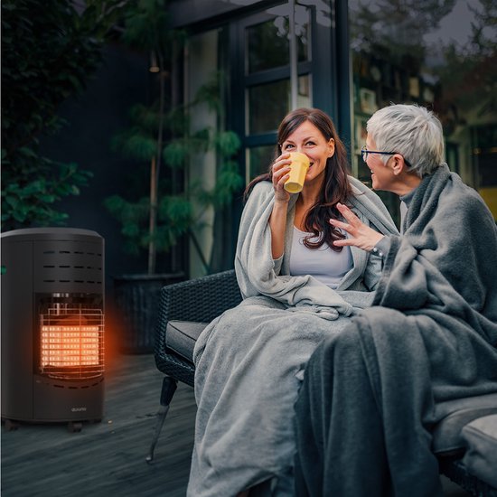 Twee vrouwen, bedekt met dekens, genieten van een warme drank op een terras naast een brandende zwarte gaskachel.
