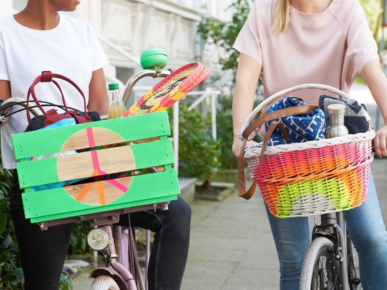 Deux femmes à vélo avec des paniers remplis d'affaires. Le panier de gauche est vert avec un signe de paix, le panier de droite est multicolore.