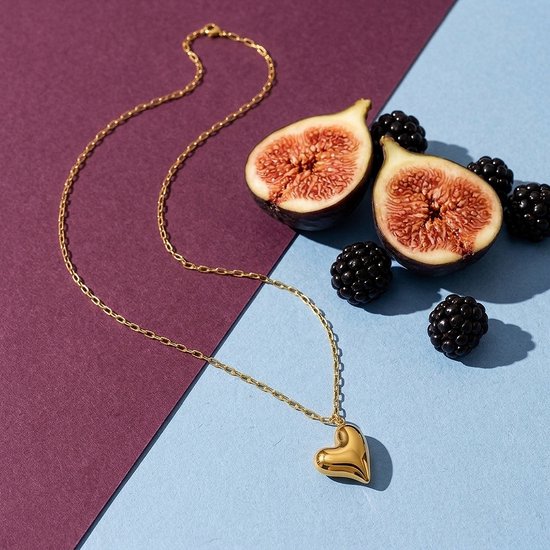 Goudkleurige ketting met hartvormige hanger, gepresenteerd naast verse vijgen en bramen op een tweekleurige achtergrond.