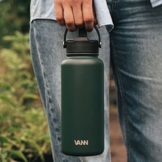 Persoon houdt een donkergroene VANN-fles vast met een zwarte dop tegen een achtergrond van natuur en jeans.