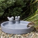 Grijze vogeldrinkschaal met twee vogelfiguren, waaraan een mus in de tuin drinkt.