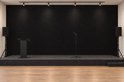 Podium met zwarte viltachterwand, houten vloer, een spreekgestoelte, een microfoonstandaard en twee zwarte luidsprekers.