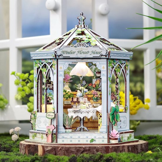 Miniatuur 'Windsor Flower House' met verlichte interieur en bloemen, staand op een boomstronk.