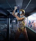 Bouwvakker met helm en masker boort in betonnen plafond met een blauwe Bosch boorhamer op een bouwplaats.
