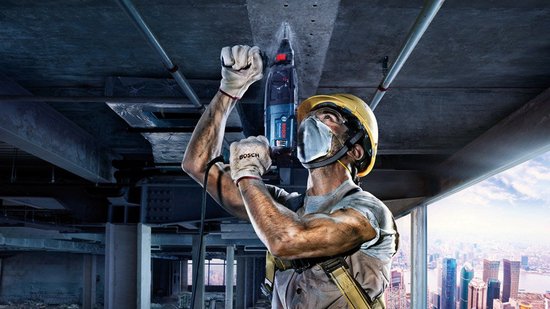Bouwvakker met helm en masker gebruikt een Bosch boorhamer aan een betonnen plafond op grote hoogte.