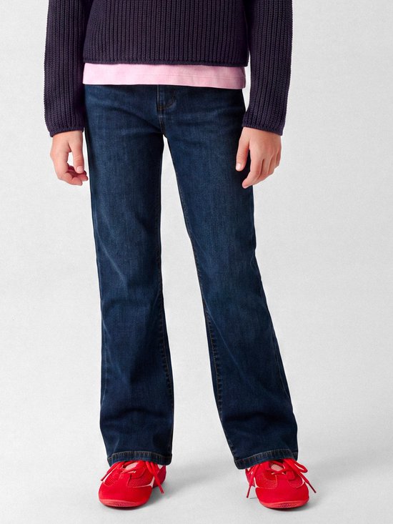 Model draagt een donkerblauwe bootcut jeans, een roze shirt en een donkerpaarse trui, gecombineerd met rode sneakers.