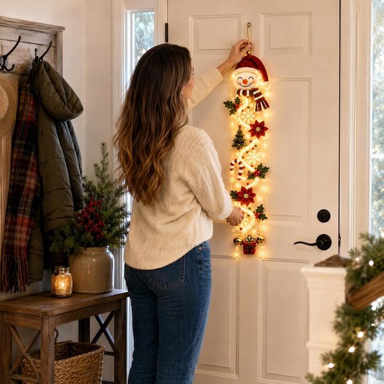 Vrouw hangt een sneeuwpop kersthanger aan een witte deur in een hal met warme sfeerverlichting.