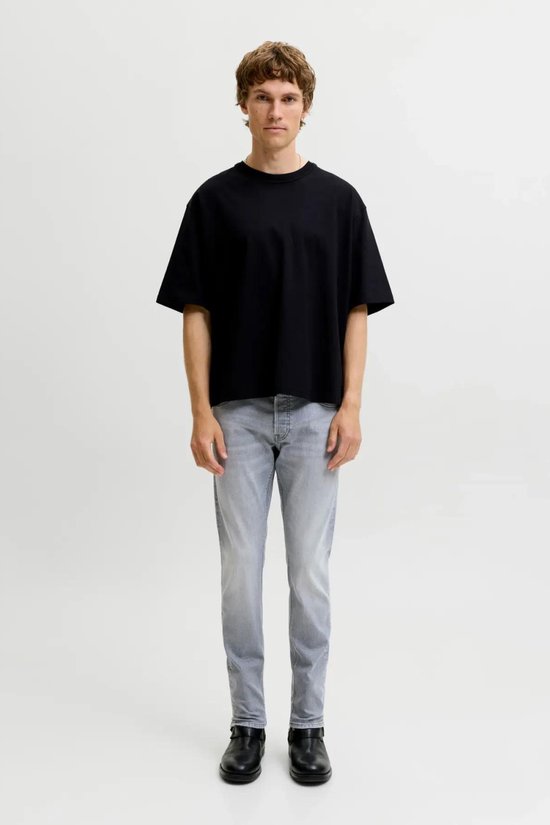 Man in zwart oversized t-shirt en grijze jeans met rechte pijpen, staand in de studio.