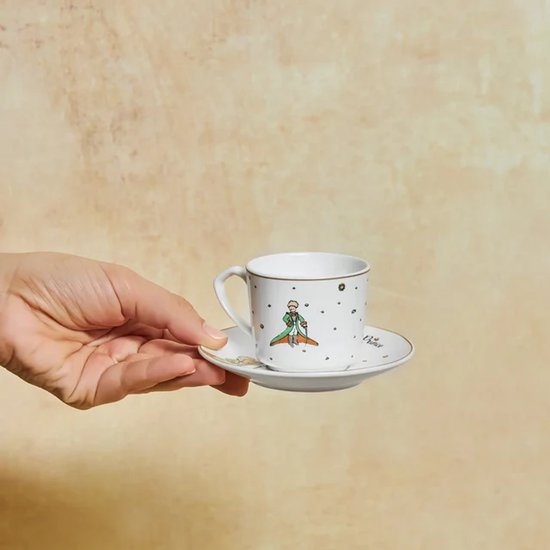 Een hand houdt een wit porseleinen espressokopje met een Le Petit Prince-illustratie en gouden rand vast.