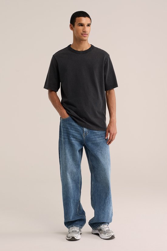 Man met handen in zakken, draagt een stonewashed spijkerbroek met losse pasvorm en een grijs T-shirt.