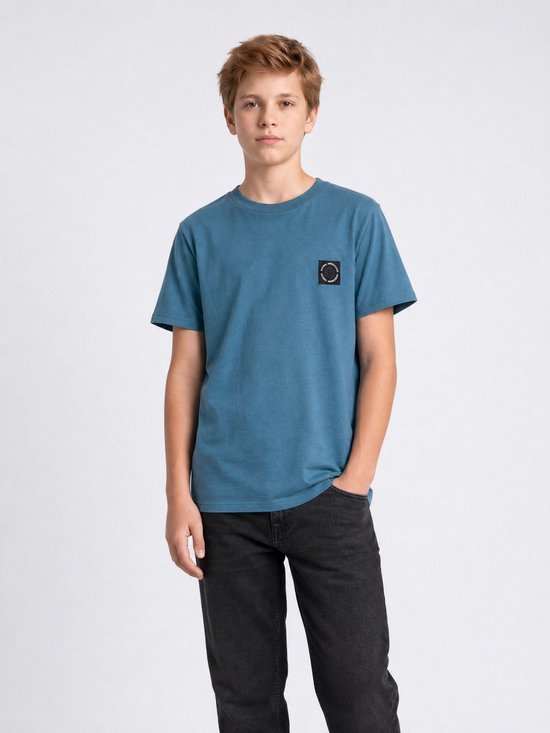 Jongen draagt een blauw katoenen T-shirt met korte mouwen en een zwart logo op de linkerborst, met handen in zijn broekzak.