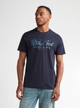 Model in een marineblauw T-shirt met 'Petrol Ind Motors Inc. Mfg. Pennsylvania' print, met spijkerbroek.