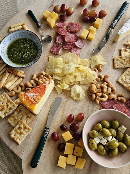 Een prachtig gedekte houten plank met diverse kazen, worstjes, druiven, olijven, crackers, chips en een dip. Een groen botermesje met sierlijk handvat ligt centraal.