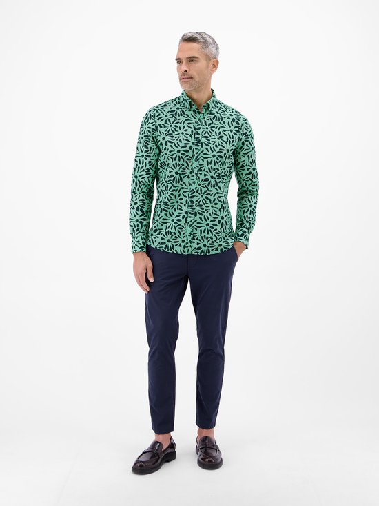 Man staand met handen in de zakken, gekleed in mintgroen overhemd met bloemenprint en marineblauwe broek.