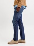 Man in een spijkerbroek, blauw overhemd en bruine jas, van de zijkant gezien.
