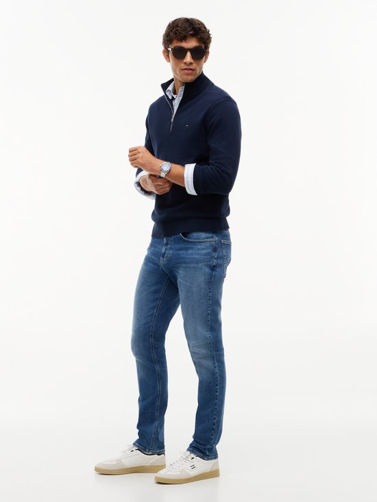 Man met zonnebril, casual shirt en donkerblauwe jeans, model staat met armen gekruist.